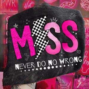 Peut inclure: T-shirt noir avec un motif graphique rose et blanc représentant le mot "MISS" avec un éclair et une couronne. Le texte "NEVER DO NO WRONG" est imprimé sous le graphique.