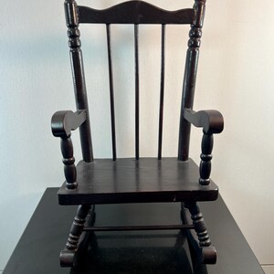 May include: A small, dark brown wooden rocking chair. The chair has a slatted back, curved armrests, and a seat. The rockers are visible at the bottom. The chair is a miniature version of a classic rocking chair.