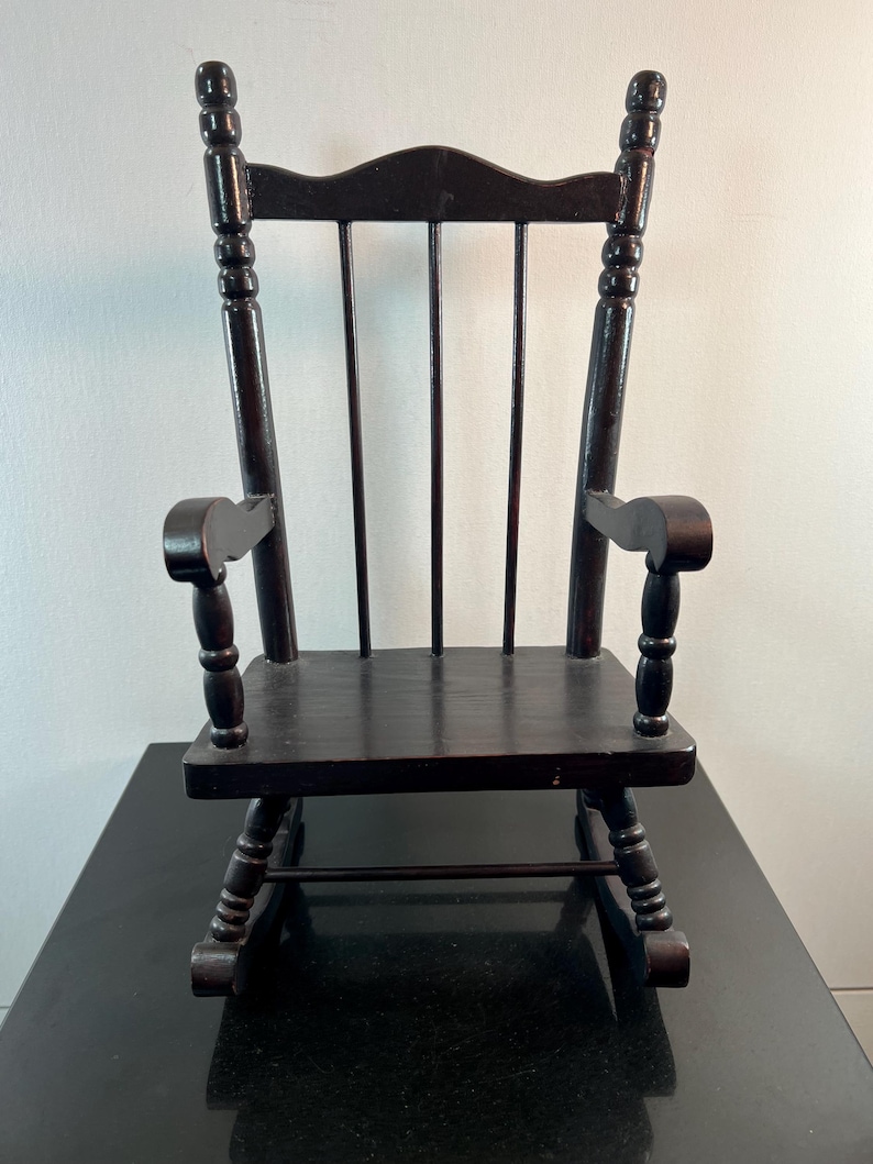 May include: A small, dark brown wooden rocking chair. The chair has a slatted back, curved armrests, and a seat. The rockers are visible at the bottom. The chair is a miniature version of a classic rocking chair.