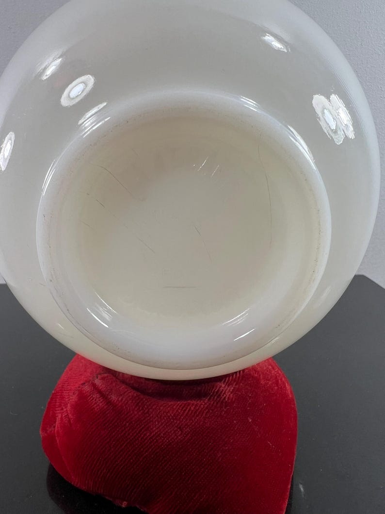 May include: A white milk glass bowl with a smooth, rounded surface. The bowl is sitting on a red velvet cushion.