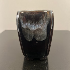 May include: A small, black ceramic pot with a white glaze. The pot has a square shape with rounded edges and four small feet.