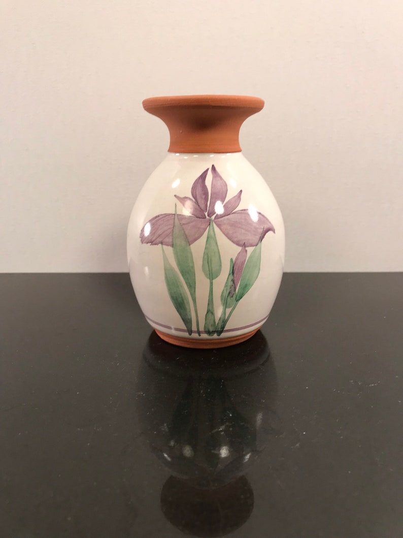 May include: A white ceramic vase with a terracotta rim, featuring a hand-painted purple iris flower design with green leaves.