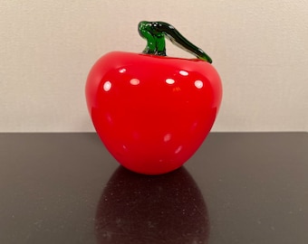 May include: A red glass apple with a green stem and leaf. The apple is sitting on a black surface.