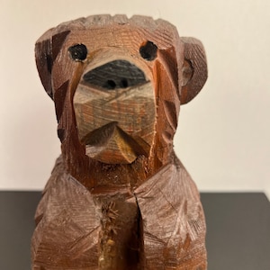 May include: A wooden carved bear figurine with a brown finish. The bear has a simple, stylized design with a rounded head and a wide, flat nose. The bear's eyes are painted black.