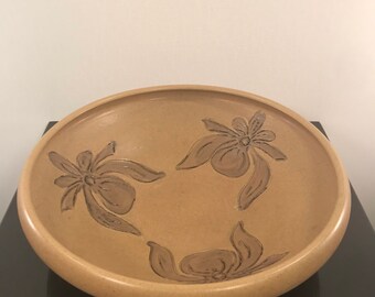 May include: A large, tan ceramic bowl with a decorative design of three stylized flowers. The bowl has a slightly rounded rim and a smooth, glazed surface.