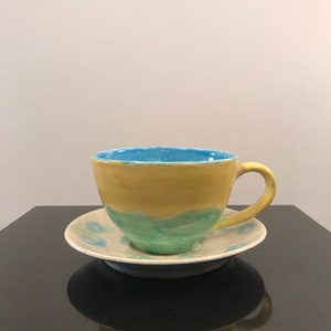 May include: A yellow, green, and blue ceramic teacup and saucer set. The cup has a rounded shape and a handle. The saucer has a simple design.