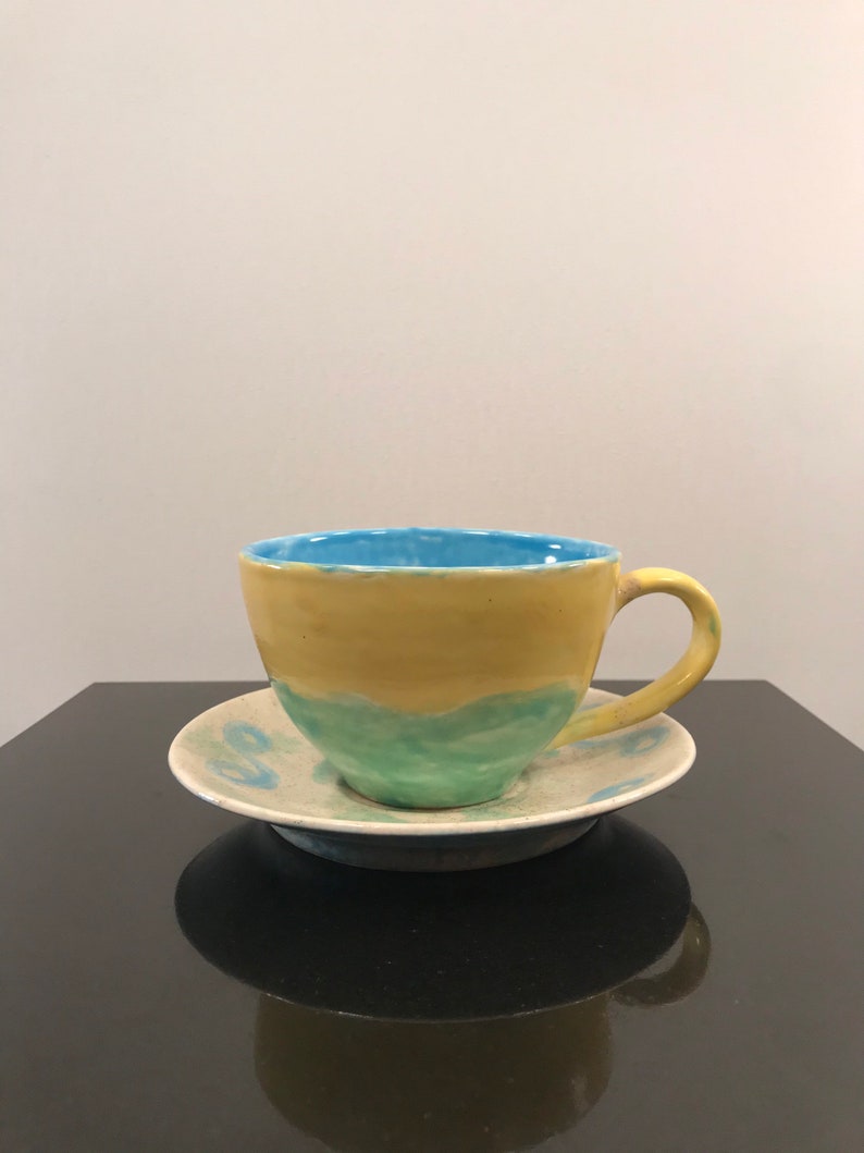 May include: A yellow, green, and blue ceramic teacup and saucer set. The cup has a rounded shape and a handle. The saucer has a simple design.