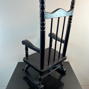 May include: A small, black wooden rocking chair with a classic design. The chair features a slatted back, curved rockers, and armrests. The chair is painted black and appears to be a miniature or child-sized piece of furniture.