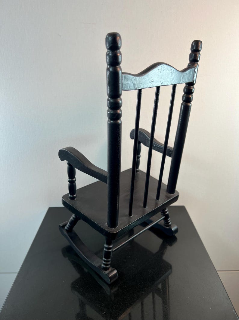 May include: A small, black wooden rocking chair with a classic design. The chair features a slatted back, curved rockers, and armrests. The chair is painted black and appears to be a miniature or child-sized piece of furniture.