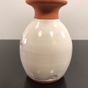 May include: A white ceramic vase with a terracotta rim and a subtle purple design. The vase has a rounded body and a slightly flared opening.