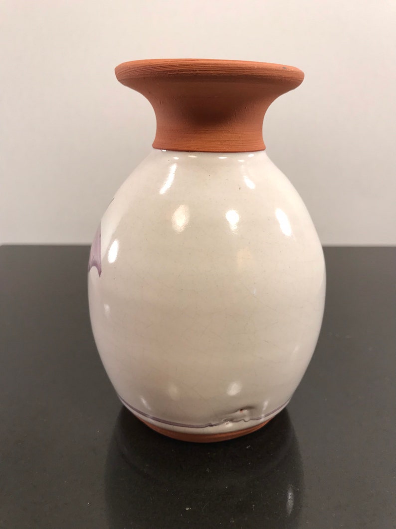 May include: A white ceramic vase with a terracotta rim and a subtle purple design. The vase has a rounded body and a slightly flared opening.