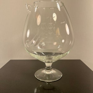 May include: A clear glass pitcher with a ship etched into the side. The pitcher has a wide base and a spout.