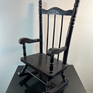 May include: A small, black wooden rocking chair with a slatted back and curved armrests. The chair has a classic design, with rounded rockers and a smooth, dark finish. It is a miniature version of a traditional rocking chair.