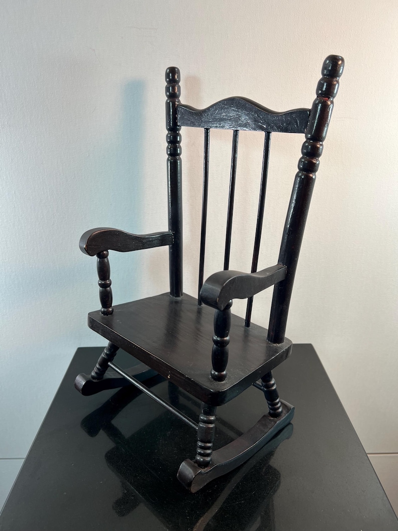 May include: A small, black wooden rocking chair with a slatted back and curved armrests. The chair has a classic design, with rounded rockers and a smooth, dark finish. It is a miniature version of a traditional rocking chair.