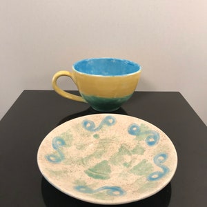 May include: A yellow, green, and blue ceramic teacup and saucer set with a swirling blue design on the saucer.