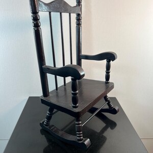 May include: A small, dark brown wooden rocking chair. The chair has a slatted back, curved armrests, and a seat. The rockers are visible at the bottom. The chair is made of wood and has a glossy finish. It is a miniature version of a classic rocking chair.