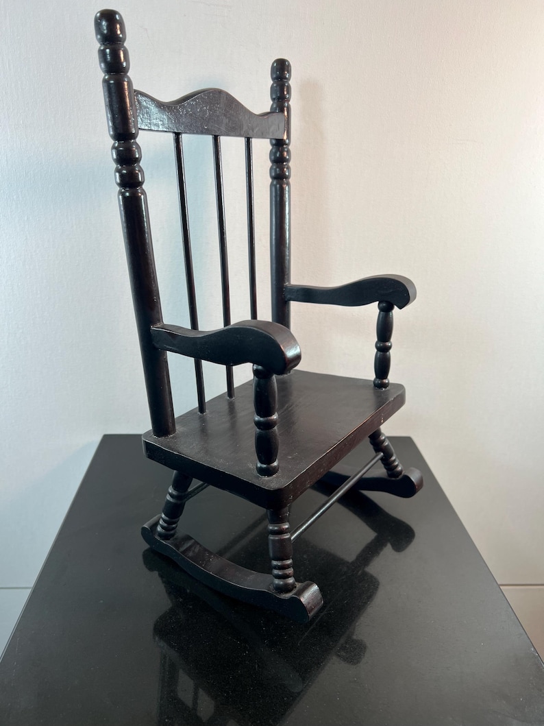 May include: A small, dark brown wooden rocking chair. The chair has a slatted back, curved armrests, and a seat. The rockers are visible at the bottom. The chair is made of wood and has a glossy finish. It is a miniature version of a classic rocking chair.