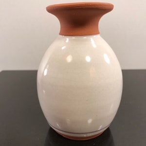 May include: A white ceramic vase with a terracotta rim and a brown stripe around the bottom. The vase has a rounded body and a slightly flared opening.
