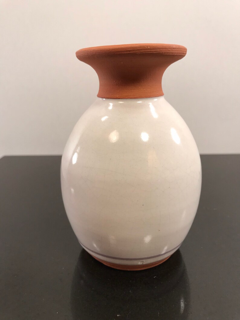 May include: A white ceramic vase with a terracotta rim and a brown stripe around the bottom. The vase has a rounded body and a slightly flared opening.