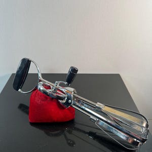 May include: A vintage chrome hand mixer with black handles and a wooden-handled beater. The mixer rests on a red velvet pouch, set against a dark surface.