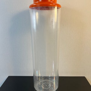 May include: A clear plastic container with an orange lid. The container is tall and cylindrical.