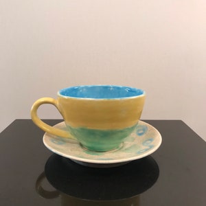 May include: A yellow and blue ceramic teacup and saucer set. The cup has a light blue interior and a yellow exterior with a green band. The saucer has a white background with blue and green swirls.