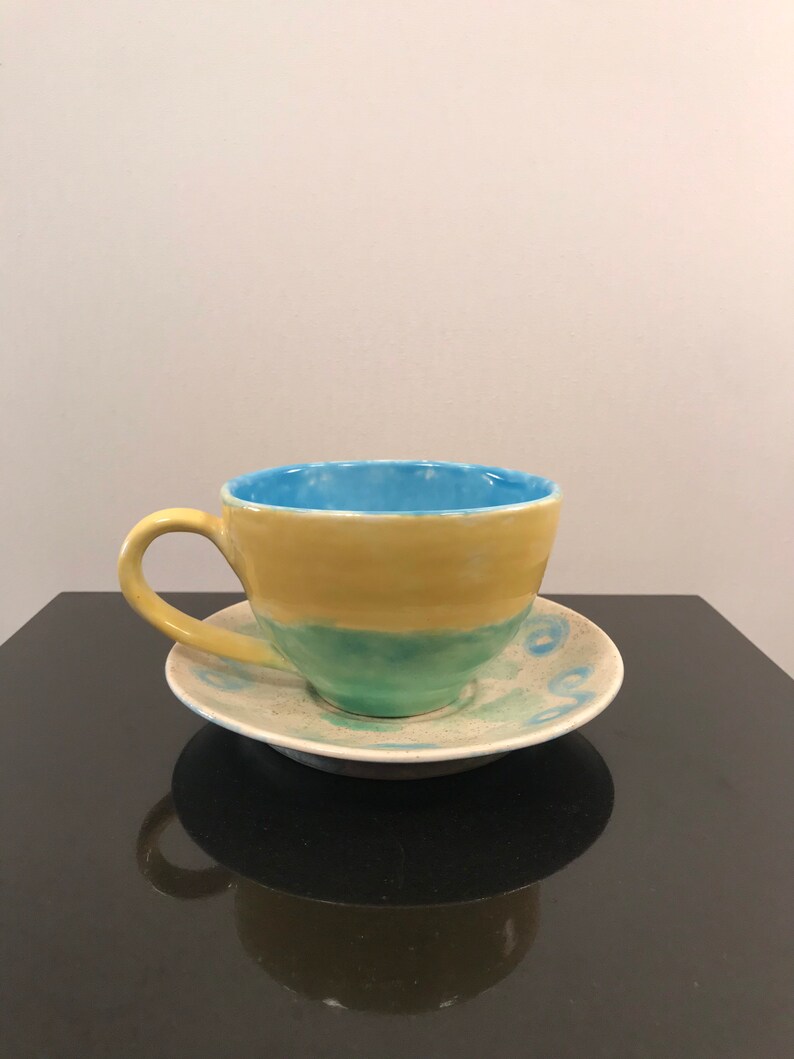May include: A yellow and blue ceramic teacup and saucer set. The cup has a light blue interior and a yellow exterior with a green band. The saucer has a white background with blue and green swirls.