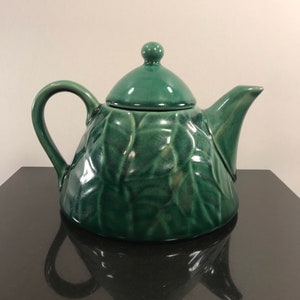 May include: A green ceramic teapot with a leaf design. The teapot has a lid with a knob on top.