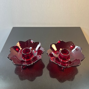 May include: A pair of red glass candle holders with a scalloped edge and three legs.