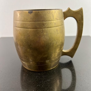 May include: A brass mug with a barrel-shaped body and a handle. The mug has a worn, aged look.