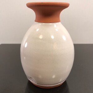 May include: A white ceramic vase with a terracotta rim and base. The vase has a rounded body and a slightly flared opening.