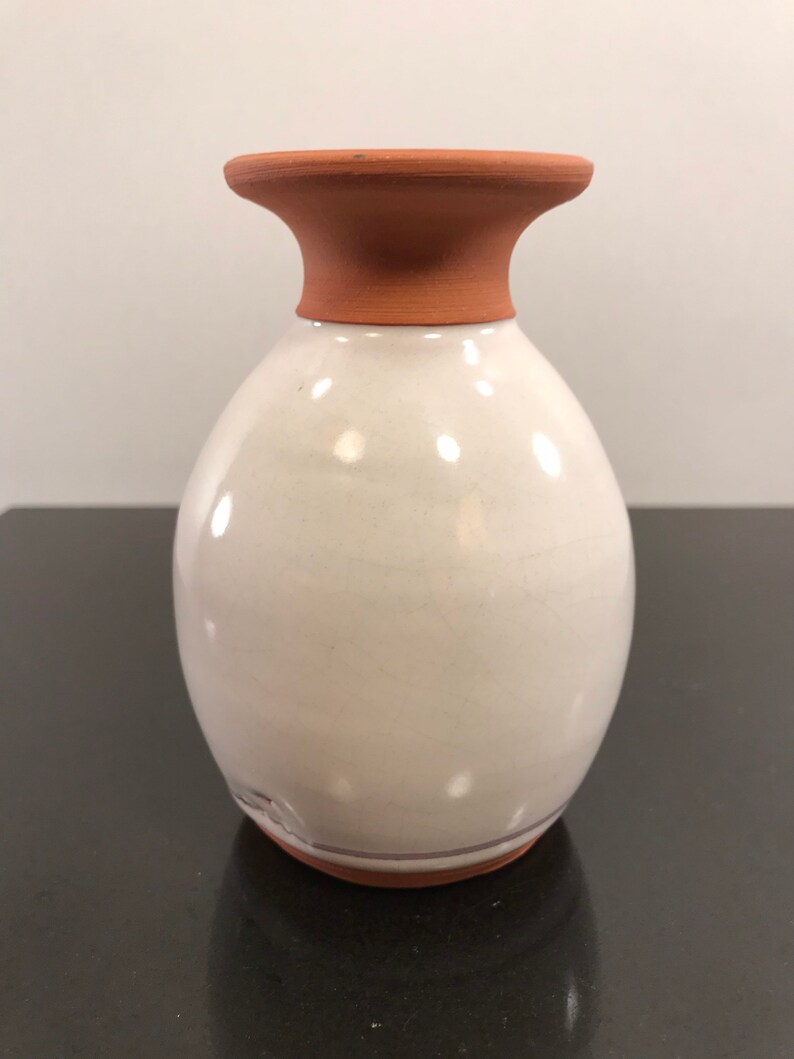 May include: A white ceramic vase with a terracotta rim and base. The vase has a rounded body and a slightly flared opening.