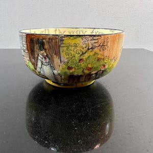 May include: A hand-painted ceramic bowl with a yellow interior and a detailed exterior scene. The bowl features a man in a top hat and a garden with red fruit. The rim has text in black. The bowl is on a black surface.