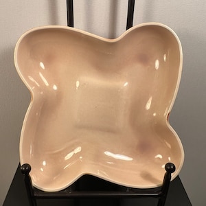 May include: A light pink ceramic bowl with an irregular, organic shape. The bowl is sitting on a black metal stand.