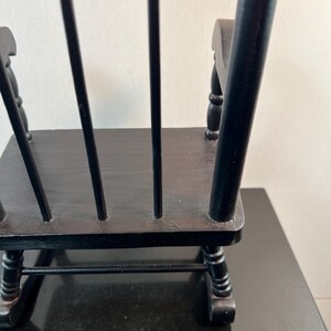 May include: A small, black wooden rocking chair with a slatted back and curved rockers. The chair is painted black and appears to be a miniature or decorative piece. The chair is on a black surface.