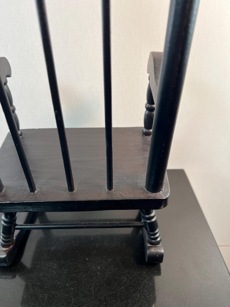 May include: A small, black wooden rocking chair with a slatted back and curved rockers. The chair is painted black and appears to be a miniature or decorative piece. The chair is on a black surface.