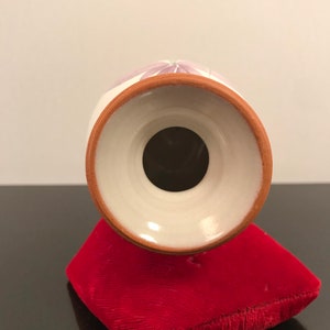 May include: White ceramic vase with a purple floral design. The vase has a round base and a wide opening. The vase is sitting on a red velvet cushion.