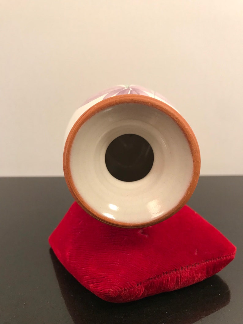 May include: White ceramic vase with a purple floral design. The vase has a round base and a wide opening. The vase is sitting on a red velvet cushion.