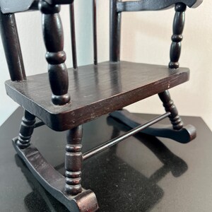 May include: A miniature wooden rocking chair in a dark brown finish. The chair features a slatted back, curved armrests, and a seat. The rockers are visible at the base. This small chair is a decorative item.