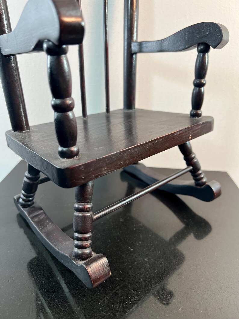 May include: A miniature wooden rocking chair in a dark brown finish. The chair features a slatted back, curved armrests, and a seat. The rockers are visible at the base. This small chair is a decorative item.