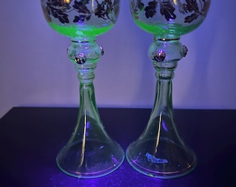 May include: Two antique green-tinted glass goblets. The bowls are clear with a gold and brown leaf design. The stems have a bulbous section with decorative accents. The goblets are displayed on a dark surface.