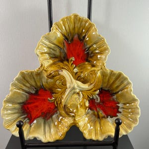 May include: A decorative, three-sectioned serving dish with a leaf-like design. The dish features a glossy finish with a color palette of yellow, red, and brown. The dish is displayed on a black metal stand, showcasing its intricate details and unique shape.