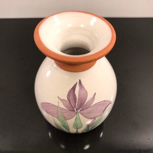 May include: A small white ceramic vase with a terracotta rim and a purple flower design.