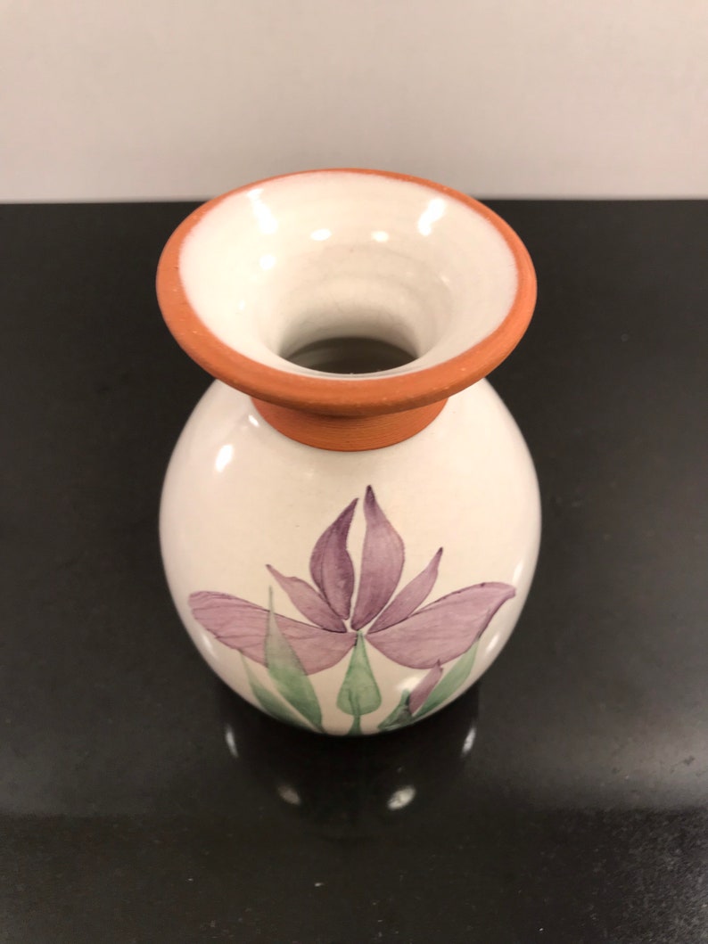 May include: A small white ceramic vase with a terracotta rim and a purple flower design.
