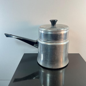 May include: A vintage aluminum double boiler with a black handle and lid knob. The pot has a cylindrical base and a smaller upper section, designed for gentle heating. The handle is angled for easy use. The pot is sitting on a black surface.