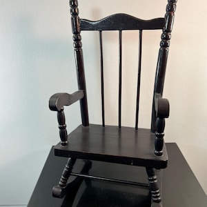 May include: A small, dark brown wooden rocking chair. The chair has a slatted back, curved armrests, and a gently curved base for rocking. The wood has a glossy finish. The chair is a miniature version of a classic rocking chair.