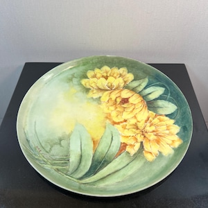 May include: A round ceramic plate featuring a hand-painted design of three large, vibrant yellow flowers with green leaves. The background is a soft, blended wash of light green and yellow tones, with a small signature visible on the lower left side.