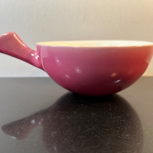 May include: A pink ceramic bowl with a handle. The bowl has a white interior and a pink exterior.