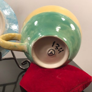 May include: A yellow, green, and blue ceramic teacup with a handle. The bottom of the cup has a white circle with the number 134 and a letter A inside.