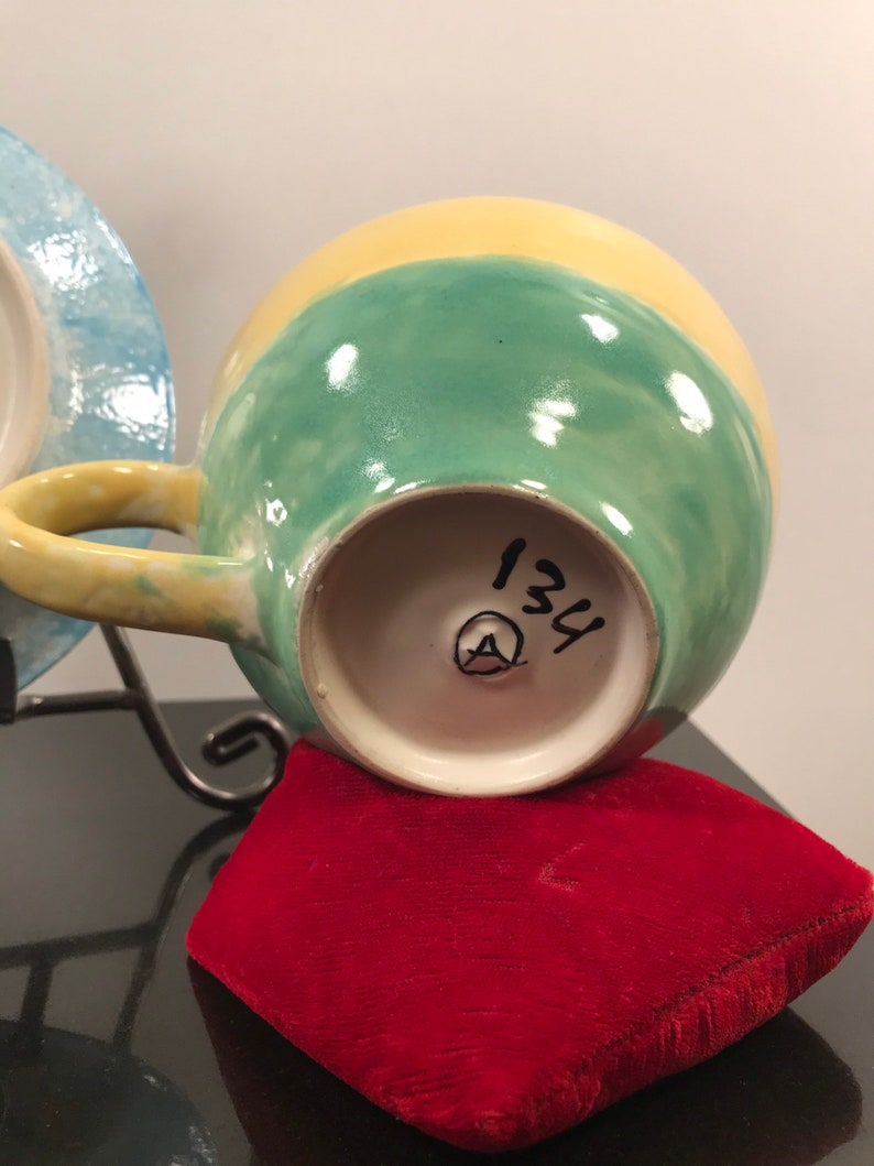 May include: A yellow, green, and blue ceramic teacup with a handle. The bottom of the cup has a white circle with the number 134 and a letter A inside.